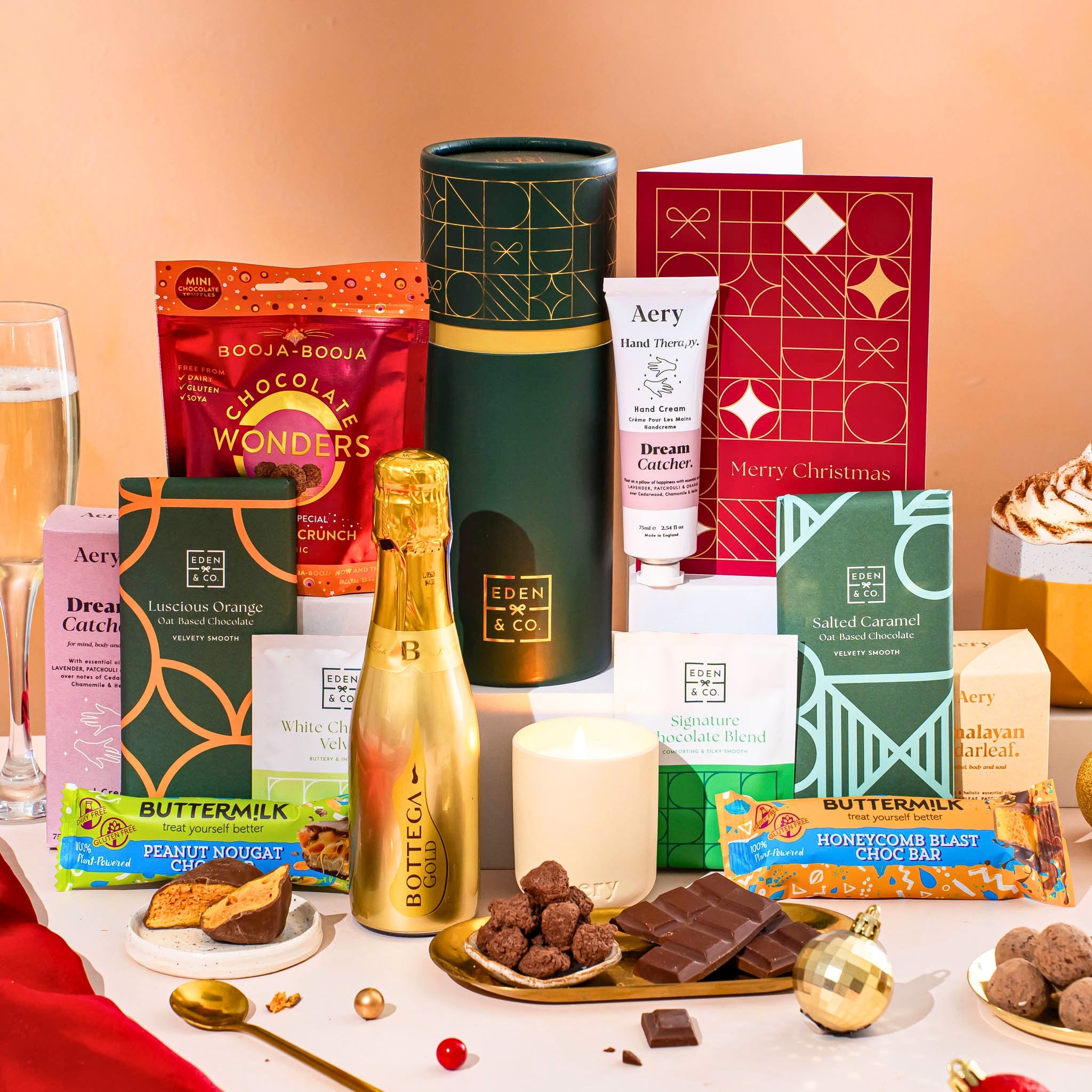 EDEN & CO. plant-based gift hamper with a mini Bottega Gold prosecco, green gift tube, “Occasion Themed” card, chocolate bars in Luscious Orange, White Chocolate Velvet, Signature Chocolate Blend and Salted Caramel, Booja-Booja Chocolate Wonders truffles, Buttermilk Peanut Nougat and Honeycomb Blast bars, chocolate truffles and honeycomb pieces on small plates, and a filled champagne glass, luxury candle and hand cream set against a warm terracotta background.