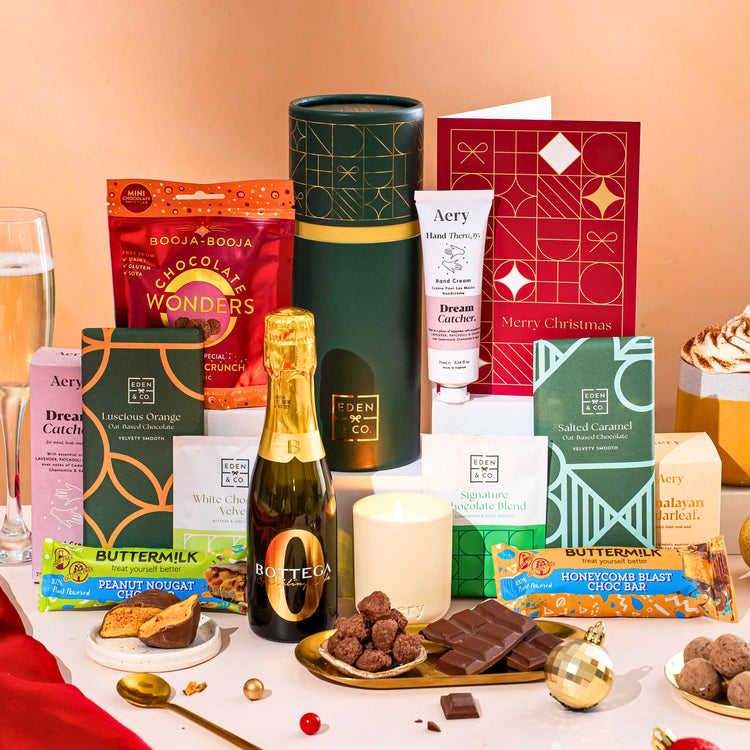 EDEN & CO. plant-based gift hamper with a mini Bottega Zero Black prosecco, green gift tube, “Occasion Themed” card, chocolate bars in Luscious Orange, White Chocolate Velvet, Signature Chocolate Blend and Salted Caramel, Booja-Booja Chocolate Wonders truffles, Buttermilk Peanut Nougat and Honeycomb Blast bars, chocolate truffles and honeycomb pieces on small plates, and a filled champagne glass,  luxury candle and hand cream set against a warm terracotta background.