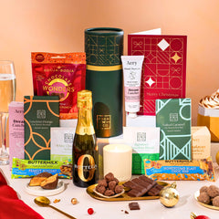 EDEN & CO. plant-based gift hamper with a mini Bottega Zero Black prosecco, green gift tube, “Occasion Themed” card, chocolate bars in Luscious Orange, White Chocolate Velvet, Signature Chocolate Blend and Salted Caramel, Booja-Booja Chocolate Wonders truffles, Buttermilk Peanut Nougat and Honeycomb Blast bars, chocolate truffles and honeycomb pieces on small plates, and a filled champagne glass,  luxury candle and hand cream set against a warm terracotta background.