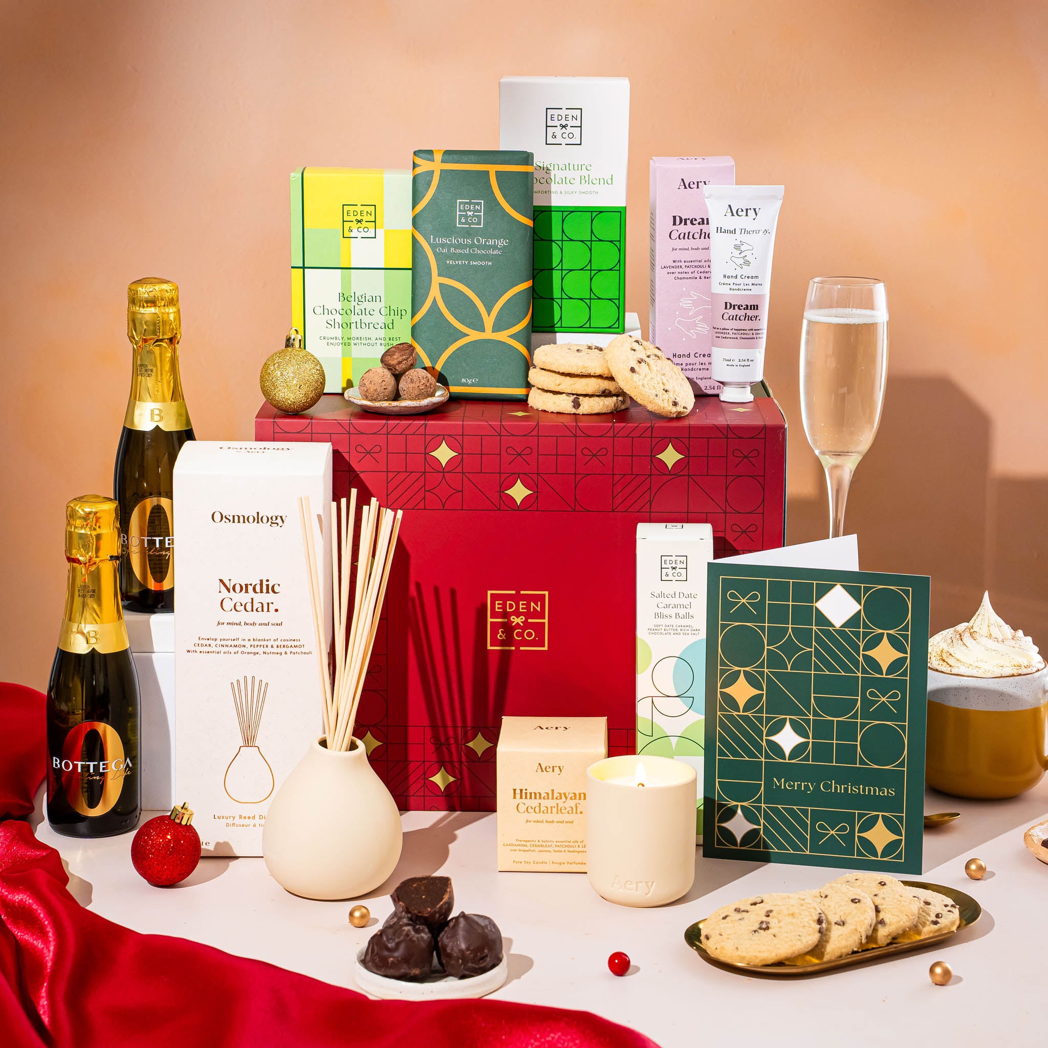 EDEN & CO. Pamper Hamper gift set with a mini Bottega Zero Black prosecco (0% alcohol-free), dark green gift cylinder, an “Occasion Themed” card, chocolate bars and truffles, a scented candle and Aery hand cream, styled with gold silk against a warm terracotta background.