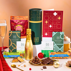 EDEN & CO. plant-based gift hamper with a mini Bottega Gold prosecco, green gift tube, “Occasion Themed” card, chocolate bars in Luscious Orange, White Chocolate Velvet, Signature Chocolate Blend and Salted Caramel, Booja-Booja Chocolate Wonders truffles, Buttermilk Peanut Nougat and Honeycomb Blast bars, chocolate truffles and honeycomb pieces on small plates, and a filled champagne glass, set against a warm terracotta background.