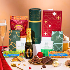 EDEN & CO. plant-based gift hamper with a mini Bottega Zero Black prosecco, green gift tube, “Occasion Themed” card, chocolate bars in Luscious Orange, White Chocolate Velvet, Signature Chocolate Blend and Salted Caramel, Booja-Booja Chocolate Wonders truffles, Buttermilk Peanut Nougat and Honeycomb Blast bars, chocolate truffles and honeycomb pieces on small plates, and a filled champagne glass, set against a warm terracotta background.