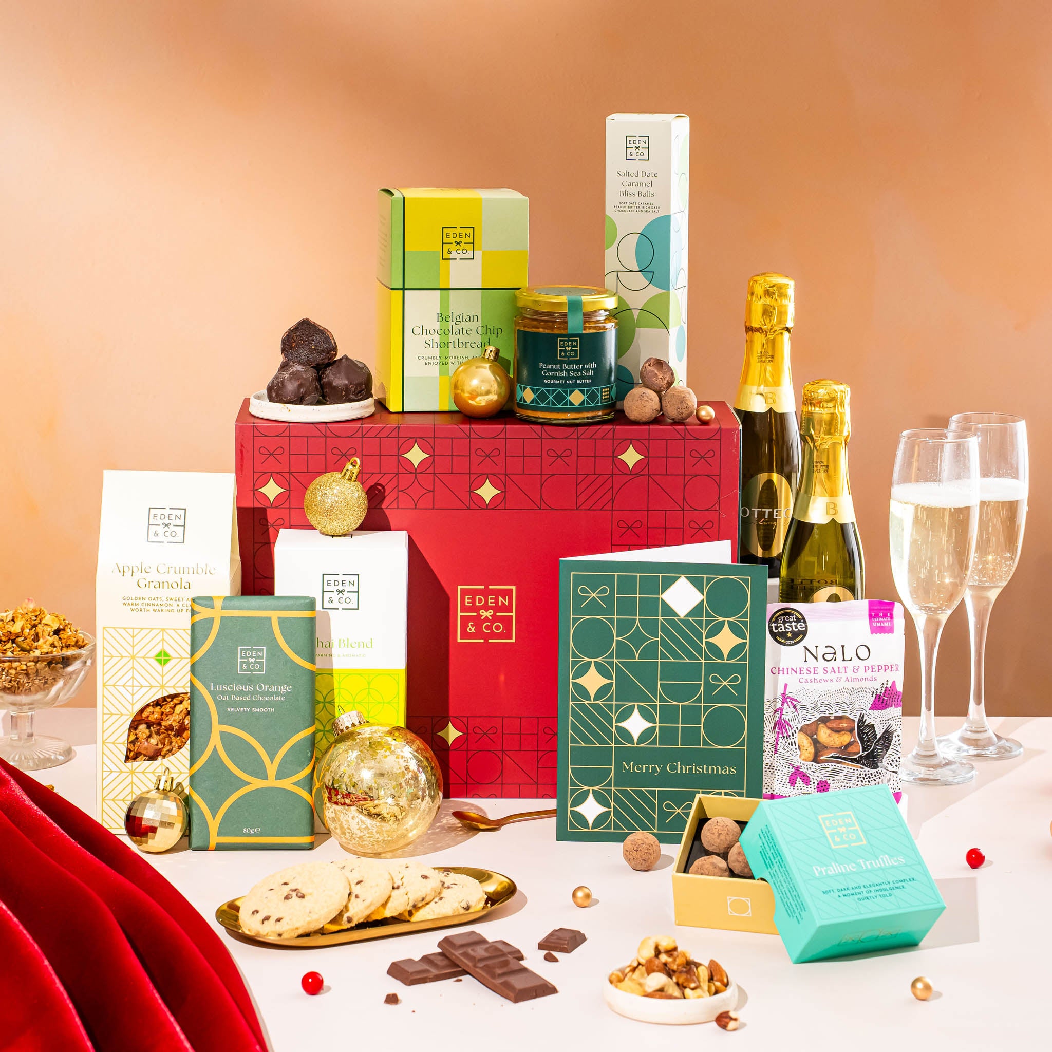 EDEN & CO. Food & Drink Hamper with a full-size Bottega Zero Black prosecco (0% alcohol-free), dark green gift cylinder, an “Occasion Themed” card, an assortment of chocolate bars, Booja-Booja Chocolate Wonders truffles, Buttermilk snack bars, a scented candle and Aery hand cream, styled with gold silk against a warm terracotta background.