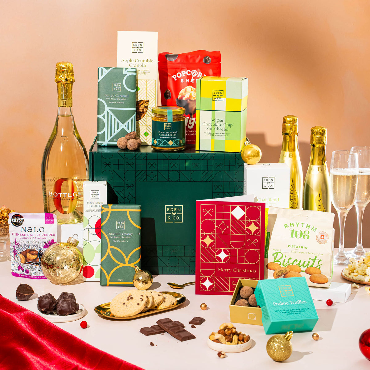 EDEN & CO. Grand Hamper gift box with a full-size Bottega Gold prosecco, dark green gift cylinder, an “Occasion Themed” card, an assortment of chocolate bars, Booja-Booja Chocolate Wonders truffles and Buttermilk snack bars, styled with gold silk against a warm terracotta background.
