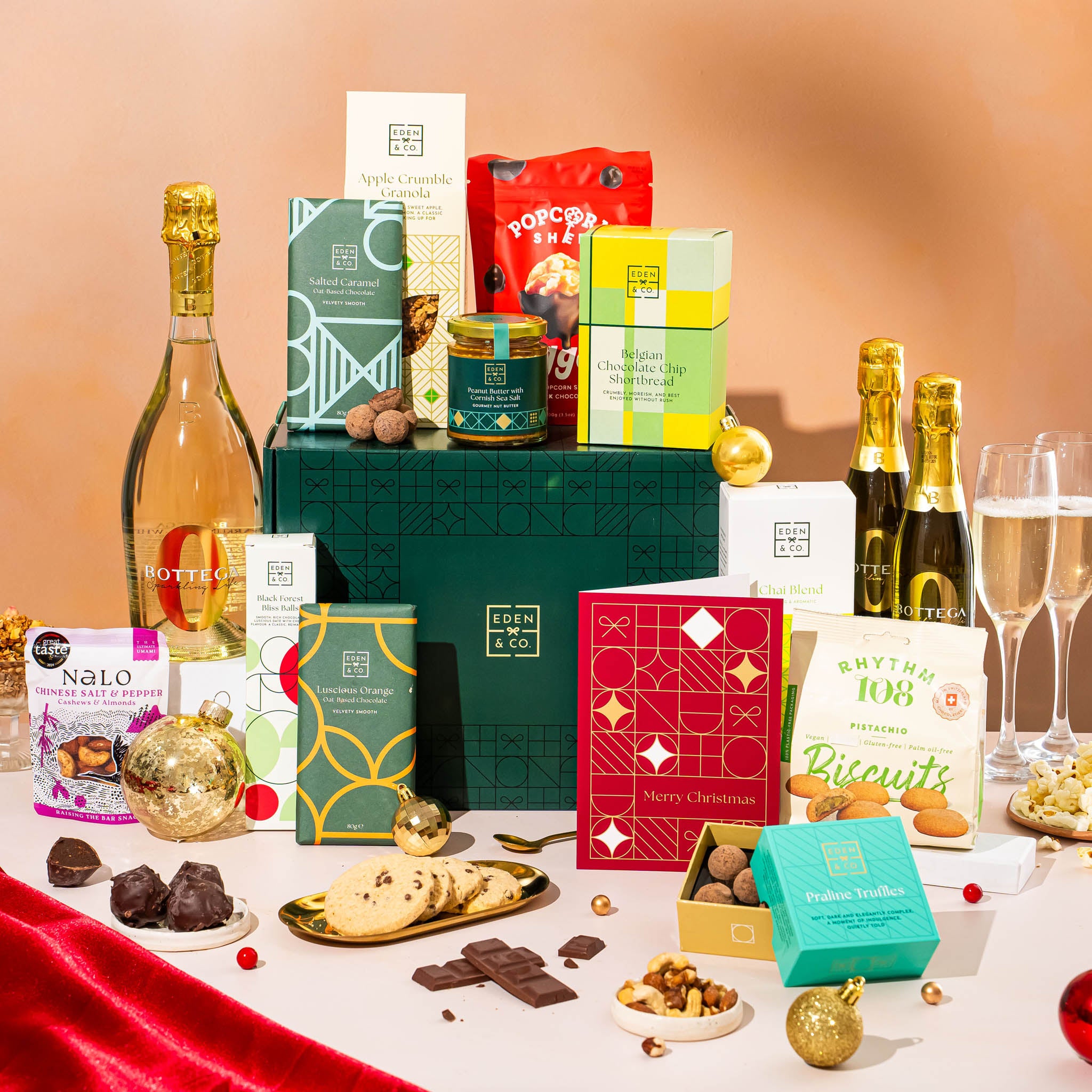 EDEN & CO. Grand Hamper gift box with a full-size Bottega Zero Black prosecco (0% alcohol-free), dark green gift cylinder, an “Occasion Themed” card, an assortment of chocolate bars, Booja-Booja Chocolate Wonders truffles and Buttermilk snack bars, styled with gold silk against a warm terracotta background.
