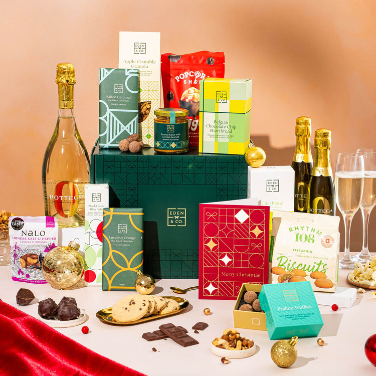 EDEN & CO. Grand Hamper gift box with a full-size Bottega Zero Black prosecco (0% alcohol-free), dark green gift cylinder, an “Occasion Themed” card, an assortment of chocolate bars, Booja-Booja Chocolate Wonders truffles and Buttermilk snack bars, styled with gold silk against a warm terracotta background.